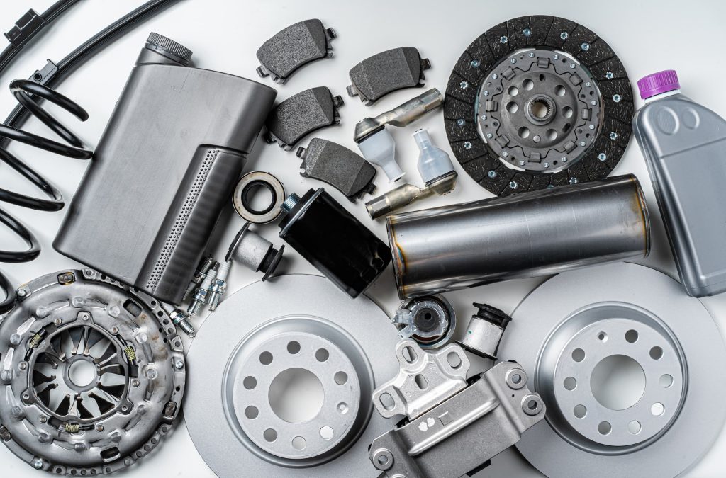 A flat lay of various new auto parts, including brake discs, shock absorbers, filters, spark plugs, and lubricants, arranged on a clean grey background.