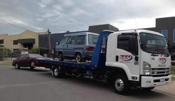 A tow truck from “MMM Auto Centre” transporting a blue van while towing a red sedan on an urban street under cloudy skies.